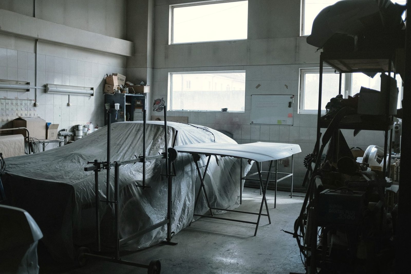 A car under a protective cover in a body shop bay with a painted panel on a stand