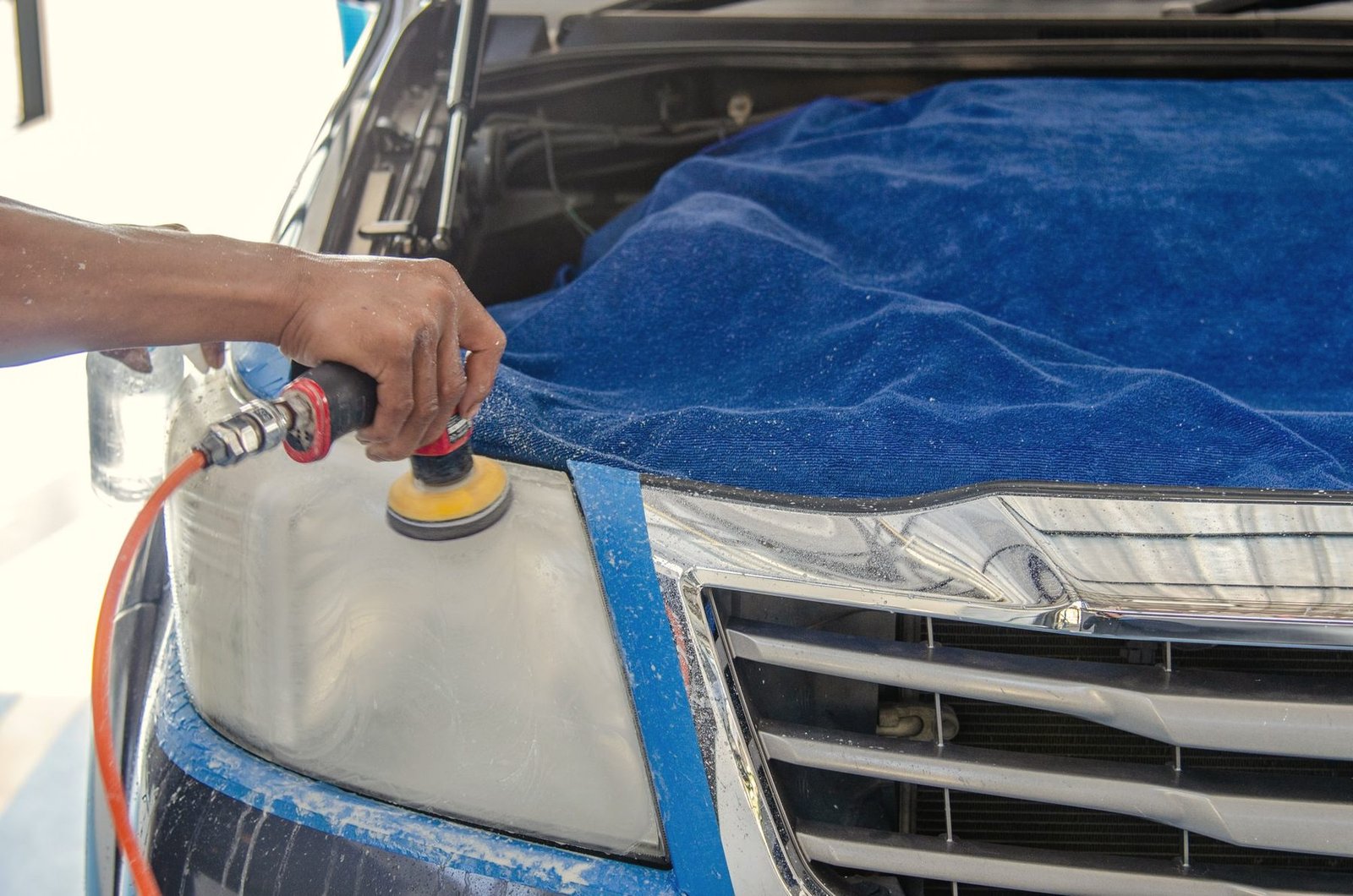 A cloudy headlight lens being polished with a small pad, the paint around it covered with blue tape and a towel
