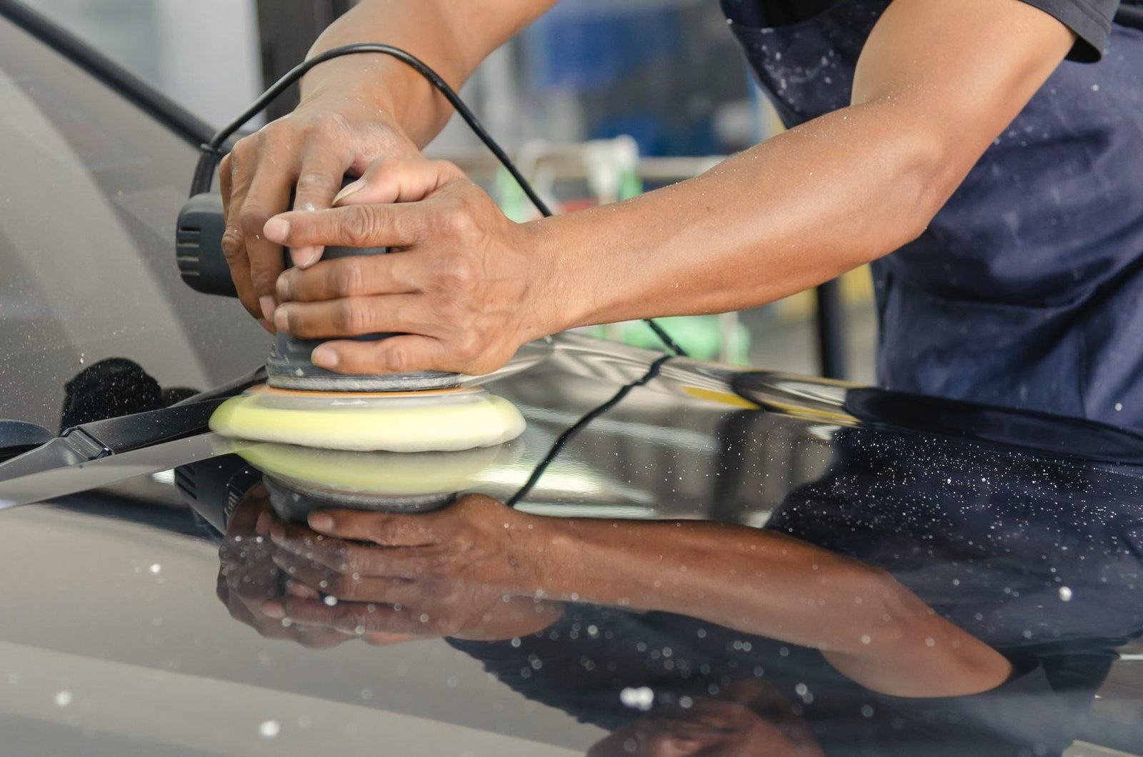 Two hands guiding a yellow polishing pad over a dark painted panel