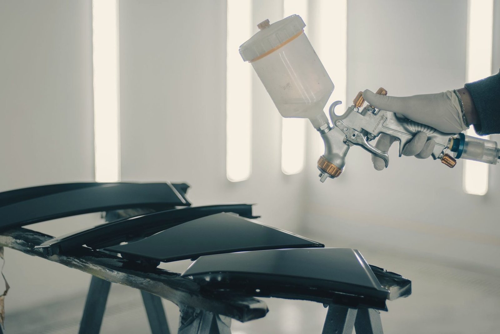 A spray gun over black trim pieces laid out on a rack in a bright paint booth